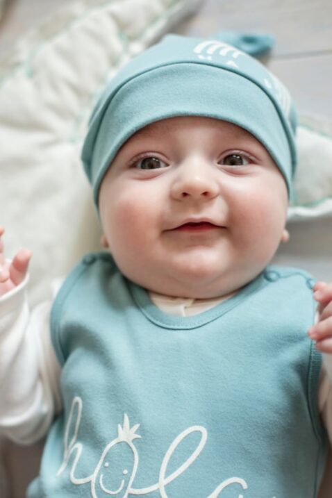 Lachendes & liegendes Baby Junge trägt grüne Mütze mit kleinem Knoten an der Spitze, grüner Strampler mit Schrifzug HOLA & weißem Wickelbody Oberteil von Pinokio - Babyphoto