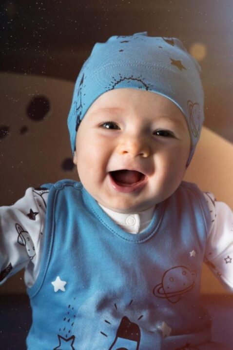 Lachender glücklicher Junge trägt blaue Baby Mütze mit Weltall Motiven Sterne Raketen Mond, blauer Weltraum Strampler & weißes Oberteil von Pinokio - Babyhoto