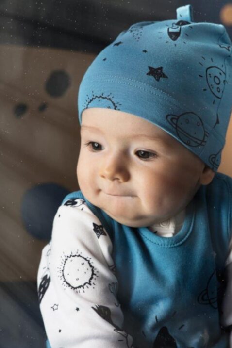 Sitzender Junge trägt blaue Baby Mütze mit Weltall Sterne Raketen Mond Motiven, blauer Strampler Weltraum & weißes Cardigan Oberteil von Pinokio - Babyhoto