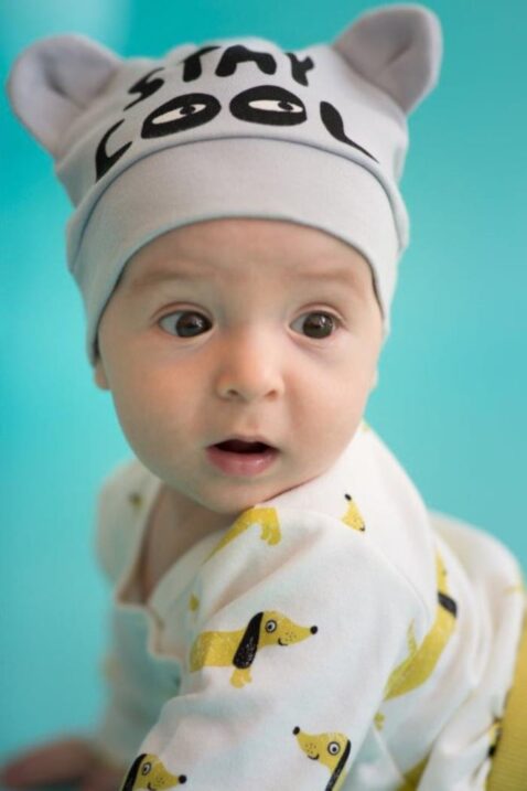 Baby staunender krabbelnder Junge trägt graue Baby Mütze mit Ohren & Schriftzug Stay Cool, weißen Wickelbody mit Hunden & gelbe Strampelhose Schlafhose mit Patch von Pinokio - Babyphoto