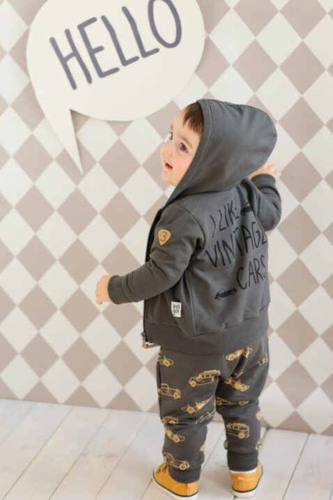 Staunender Junge trägt graue Baby Pumphose Haremshose Schlupfhose mit Autos Oldtimer Sportwagen Rennwagen Motiven & Tasche, dunkelgraue Babyjacke Kapuzen-Sweatjacke Autos, braune Babyschuhe von Pinokio - Babyphoto Rückenansicht