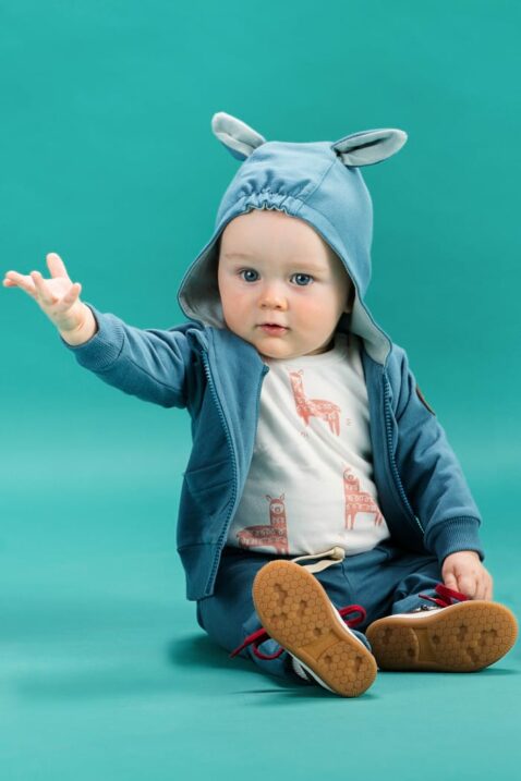 Sitzender Junge trägt weißen Baby Body ärmellos ohne Ärmel mit Lama Motiven - Blaue Baby Sweatjacke Kapuzenjacke langarm mit Lama Ohren - Blaue Babyhose mit Taschen & Happy Lama Patch von Pinokio - Babyphoto