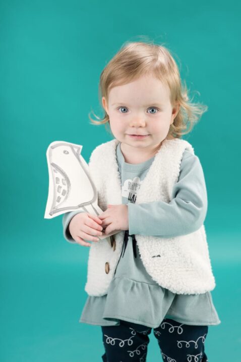 Stehendes lächelndes Baby Mädchen trägt Tunika Kleid Langarmshirt in Kleidchenoptik mit Lama Motiv - weiße gefütterte Weste ohne Ärmel mit Holz Knöpfe im Lama Wolle Design - blaue Leggings Schlupfhose mit Schleifen Lama Wolle Design von Pinokio - Babyphoto