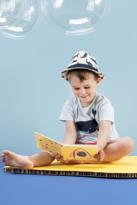 Sitzender Junge liest ein Buch & trägt hellblaues Baby Basic T-Shirt Babyshirt mit Wal Meer Motiv - Sommer rote kurze Shorts Hose aus Baumwolle mit Patch Save the world & elastischen Gummizugbund mit Kordel - Blau weißer Baby Sommerhut - Kinderphoto