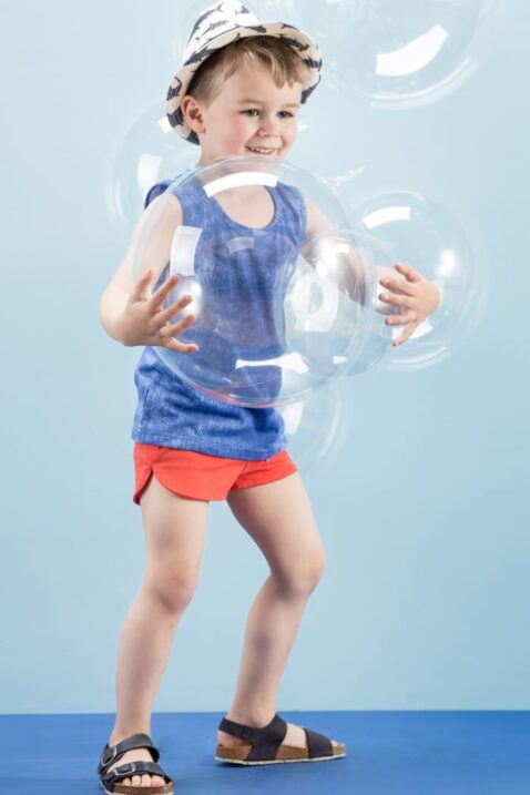 Lachender Junge trägt rote kurze Shorts Babyhose mit Patch - Blaues ärmelloses T-Shirt Tank-Top Trägertop Achselshirt im Jeans-Look - Blau-weißer Strohhut & blaue Kindersandalen von Pinokio - Kinderphoto