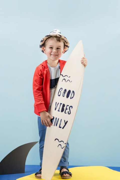 Stehender Junge mit Surfbrett trägt blaues Babyshirt kurzarm T-Shirt mit Wal Motiv - Rote lange Sweatjacke mit Kapuze - Lange blaue Sweathose Babyhose für den Sommer - Heller Strohhut & Kindersandalen marineblau - Kinderphoto