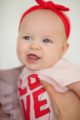 Lachendes Baby Mädchen Kind auf dem Arm der Mutter trägt rosa Body Wickelbody kurzarm kurze Ärmel im LOVE & LIEBE 3D Buchstaben Design Oberteil - Rotes Stirnband mit Schleife Erstlingsoutfit von Pinokio - Babyphoto