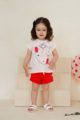 Stehendes lachendes Mädchen Kind trägt kurze rote Shorts Hose mit Rüschen & Kordel - Rosa kurzarm Sommer T-Shirt mit Erdbeer-Motiv Rundhals - Weiße Badesandalen von Pinokio - Kinderphoto