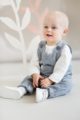 Lachender sitzender Baby Junge trägt graue Baby Latzhose Trägerhose mit Tiger Patch, kleiner Brusttasche & Beinumschlag - Weißes Langarmshirt mit Zebra Motiv - Weiße Klettverschluss Babyschuhe von Pinokio - Babyphoto