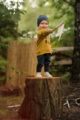 Lächelnder Junge auf einem Baumstamm trägt gelbes Baby Sweatshirt Oversize mit Zierknöpfe & Fuchs Motiv - Blaue Leggings mit Eichel & Blatt - Dunkelblaue Babymütze mit Patch von Pinokio - Kinderphoto Babyphoto