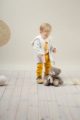 Stehender Junge trägt senfgelbe Babyhose mit Knöpfe - Weißen Papagei Babybody langarm - Plüschjacke mit Reißverschluss & Kapuze in Beige von Pinokio - Kinderphoto