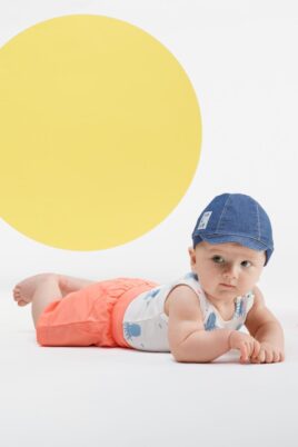 Baby Junge trägt weißen Babybody ohne Arm mit Tintenfische - Kurze Hose Shorts mit Patch & Kordel in Koralle - Blaue Schirmmütze im Jeans Look von Pinokio - Babyphoto