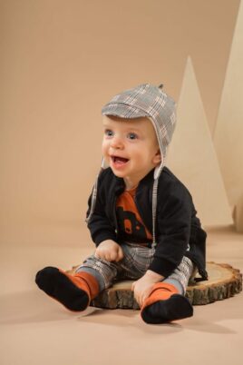 Sitzender Junge trägt graue Baby Leggings Sweathose Schottenkaro - Orange Braun Bär Wickelbody - Schwarze Sweatjacke - Pilotenmütze Schottenmuster von Pinokio - Babyphoto