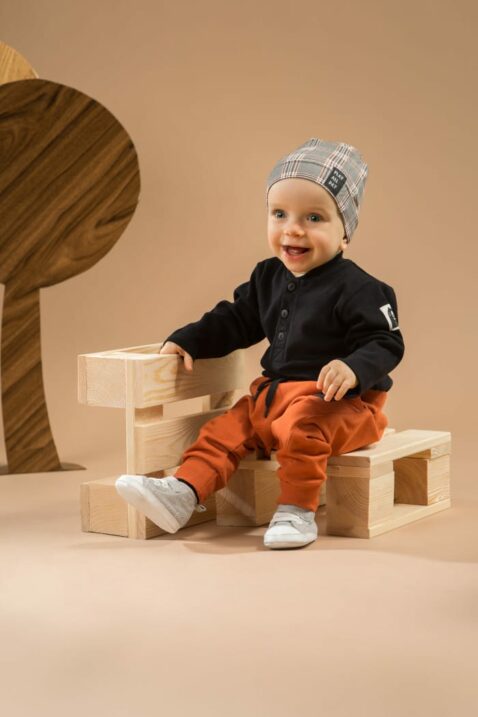 Sitzender Junge trägt rost braune Pumphose mit Taschen - Schwarzer Baby Body in Polo-Optik Patch & Knöpfe - Mütze im Schottenmuster grau Southdown Grey von Pinokio - Babyphoto