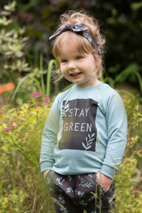 Stehendes Kinder Mädchen trägt Baby grünes Sweatshirt langarm STAY GREEN - Haremshose mit Blätter in Braun gemustert - Stirnband mit Schleife von Pinokio - Babyphoto Kinderphoto