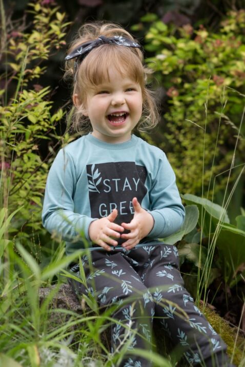 Lachendes Mädchen trägt Baby Kinder türkis grünes Langarm Shirt STAY GREEN Statement - Braunes Stirnband & Pumphose mit Zweige & Blätter Muster mit Kordel von Pinokio - Babyphoto Kinderphoto