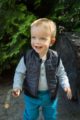 Lachender Junge trägt blau gestreiften Baby Polo Langarmbody - Schwarze Steppweste mit Auto Patch - Türkise Babyhose mit Taschen & Knöpfe von Pinokio - Babyphoto Kinderphoto