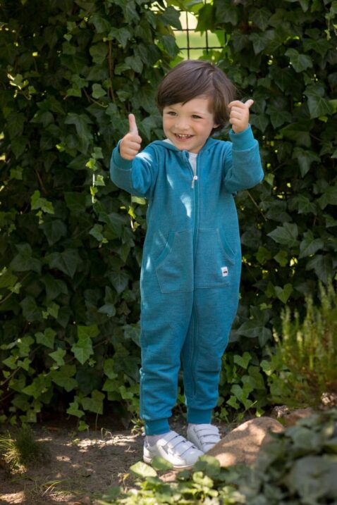 Stehender Junge trägt blauen Kinder Baby Strampler ohne Fuß, mit Kapuze, Reißverschluss, Kängurutasche & Auto Patch aus hochwertiger Baumwolle von Pinokio - Babyphoto Kinderphoto