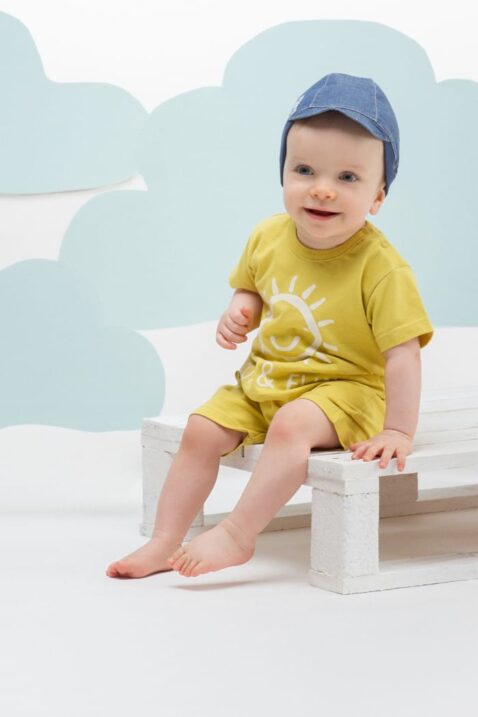 Lächelnder Junge trägt Kindermütze mit Schirm in Jeansblau - Hochwertiger Baby Romper Spieler kurzer Overall mit Sonne Gelb von Pinokio - Babyphoto Kinderphoto