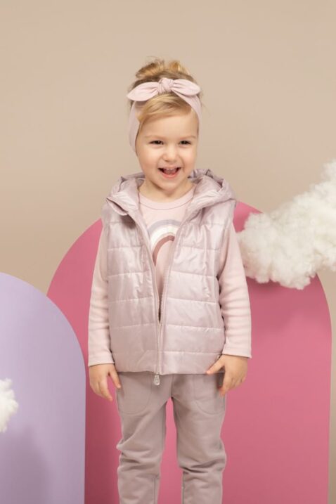 Mädchen trägt graue Babyhose mit großen Taschen & Kordel - Rosa Kinderweste Stepp Optik mit Kapuze & Taschen - Stirnband mit Schleife Basic rosa unifarben - Rosa Langarmshirt Oberteil mit Regenbogen von Pinokio - Stehendes Kind Kinderphoto