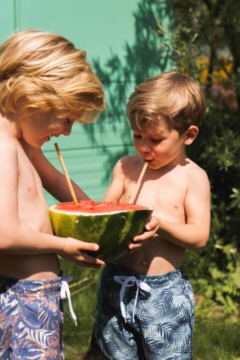 Dunkelgrüne kurze Jungen Badeshorts mit Palmen Blattmuster im All-Over-Print, Taschen, Komfortbund mit Kordel für Babys & Kleinkinder von Koko Noko - Kinderfoto Kinderbademode 2 Jungen im Garten mit süßer Swimshorts trinken aus einer Melone